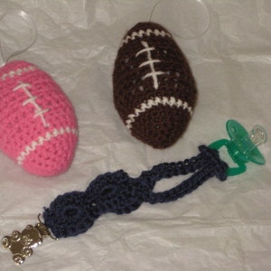 May include: Two crocheted footballs, one pink and one brown, with white stitching. A green pacifier with a blue crocheted strap and a teddy bear charm.