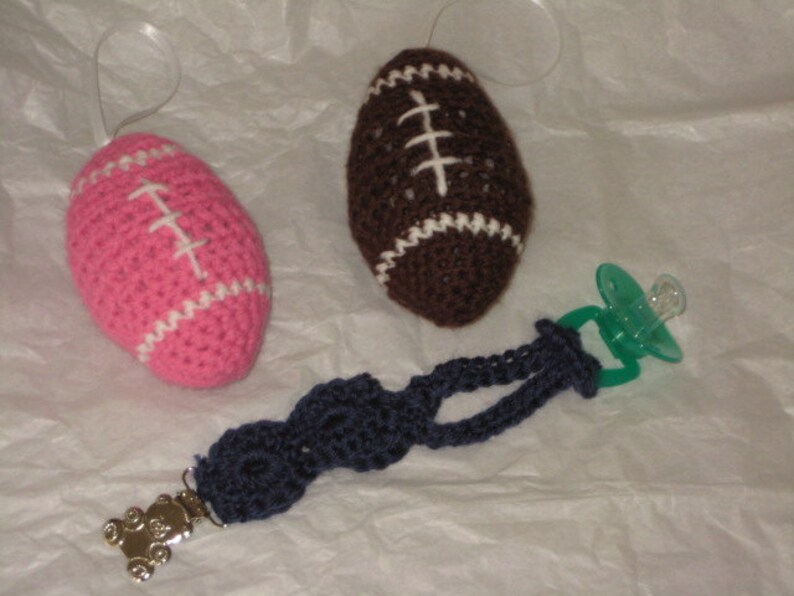 May include: Two crocheted footballs, one pink and one brown, with white stitching. A green pacifier with a blue crocheted strap and a teddy bear charm.