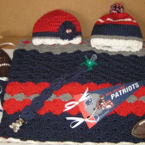 May include: A crocheted baby blanket in navy blue, red, and gray with a white trim. The blanket features a football and a pacifier. There is also a crocheted hat in red, white, and blue with a white trim.