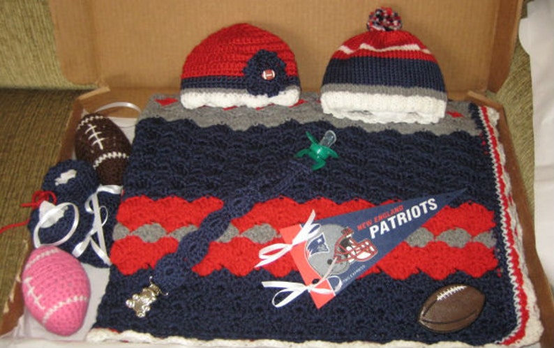 May include: A crocheted baby blanket in navy blue, red, and gray with a white trim. The blanket features a football and a pacifier. There is also a crocheted hat in red, white, and blue with a white trim.