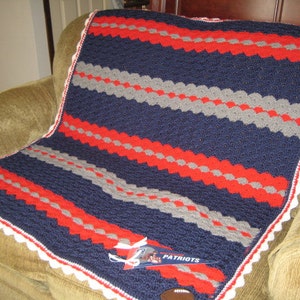 May include: A blue, red, and gray crocheted blanket with a white crocheted border. The blanket has a striped pattern and is folded on a brown couch. The blanket has a white tag with the word "Patriots" on it.