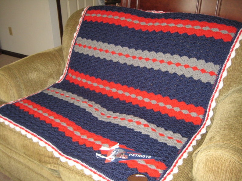 May include: A blue, red, and gray crocheted blanket with a white crocheted border. The blanket has a striped pattern and is folded on a brown couch. The blanket has a white tag with the word "Patriots" on it.