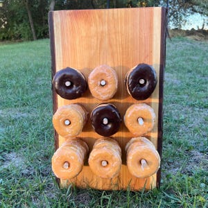 May include: A wooden donut display stand holds nine glazed donuts, some with chocolate frosting. The donuts are arranged in three rows of three, with a wooden dowel in each donut hole. The stand is made of light-colored wood.