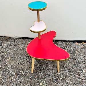 May include: A retro-style plant stand with three tiers. The top tier is a round, turquoise surface with a gold trim. The middle tier is a pink heart shape with gold trim. The bottom tier is a red boomerang shape with gold trim. The stand has wooden legs.