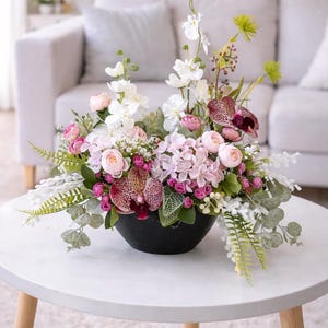 Könnte beinhalten: Blumenarrangement in einer schwarzen Schale mit rosa, weißen und burgunderfarbenen Blumen, darunter Orchideen und Rosen. Die Anordnung steht auf einem weißen Tisch mit Holzbeinen.