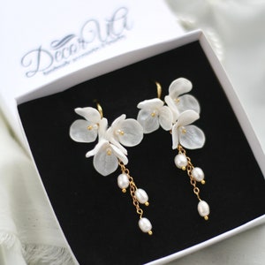 Könnte beinhalten: Weiße Blumenohrringe mit goldenen Akzenten und hängenden Perlen, präsentiert in einer weißen Schachtel mit schwarzem Innenraum. Die Ohrringe haben zarte, blütenblattartige Designs, ideal für besondere Anlässe.