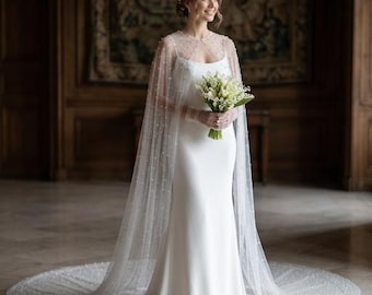 May include: A white wedding dress with a floor-length skirt and a sheer cape adorned with small, scattered pearls. The bride holds a bouquet of white flowers. The dress has a simple, elegant design.