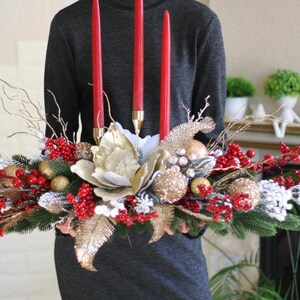 May include: A festive centerpiece featuring three red candles, gold and silver ornaments, red berries, and faux greenery. The arrangement includes a large, cream-colored flower and gold accents, perfect for holiday decor.
