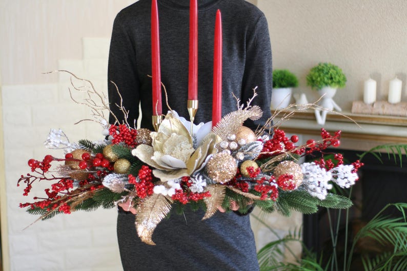 May include: A festive centerpiece featuring three red candles, gold and silver ornaments, red berries, and faux greenery. The arrangement includes a large, cream-colored flower and gold accents, perfect for holiday decor.