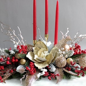 May include: A festive centerpiece with three red candles, gold ornaments, and faux berries. The arrangement includes a large gold flower and silver branches, creating a holiday display.