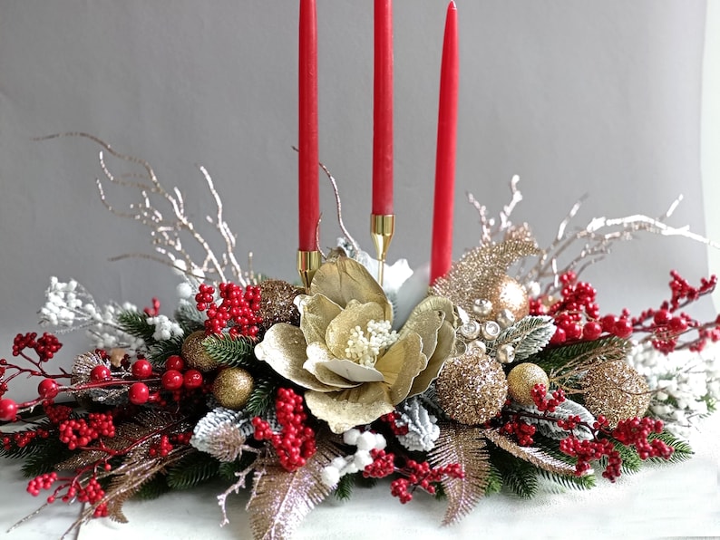 May include: A festive centerpiece with three red candles, gold ornaments, and faux berries. The arrangement includes a large gold flower and silver branches, creating a holiday display.