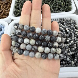 May include: A hand displays several strands of round, gray and black patterned gemstone beads. The beads are a mix of light and dark gray, with a marbled appearance. The background shows more beads in white plastic containers.