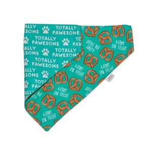May include: A turquoise blue bandana with a white paw print and the text "Totally Pawsome" repeated. The bandana is also covered in a pattern of brown pretzel shapes with the text "Little Bit Salty" repeated.