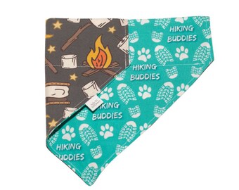May include: A turquoise and gray dog bandana with a campfire and marshmallow print on one side and a paw print and "Hiking Buddies" text on the other side.