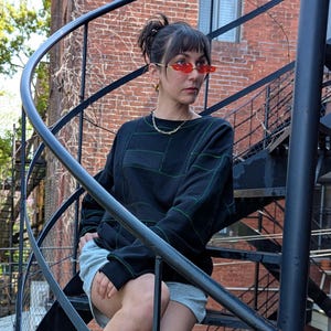 May include: A black long-sleeve sweatshirt with green grid-like stitching, light grey shorts, white crew socks, and white high-top canvas trainers. The outfit is accessorised with red-tinted oval sunglasses, a gold chain necklace, and small gold hoop earrings.