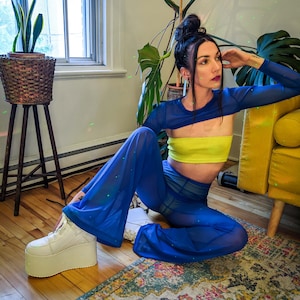 May include: A person wearing a blue mesh pantsuit with a yellow crop top. The pantsuit is sheer and has a loose fit. The person is sitting on a rug with a white platform shoe.
