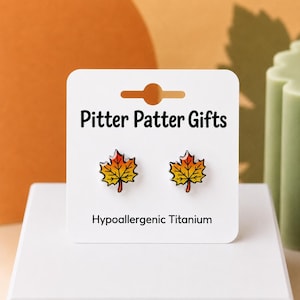 May include: A pair of stud earrings shaped like maple leaves with a gradient of red, orange, and yellow enamel. The earrings are attached to a white display card labeled Pitter Patter Gifts and Hypoallergenic Titanium with a cutout at the top.