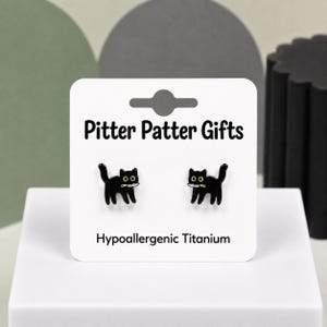 May include: A pair of black cat stud earrings with white and gold accents, each depicting a cat holding a small knife in its mouth. The earrings are displayed on a white card labeled Pitter Patter Gifts and Hypoallergenic Titanium.