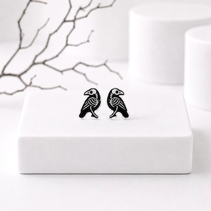 May include: A pair of stud earrings featuring a black and white graphic design of a bird skeleton. The bird is depicted in profile with visible rib cage and skull details, rendered in a high-contrast black enamel finish on a white background.