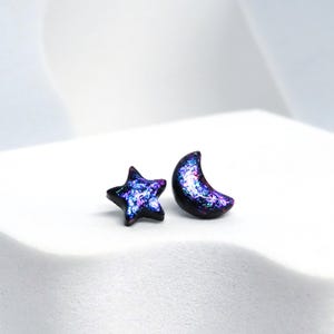 May include: A pair of celestial-themed earrings. One earring is a star shape, and the other is a crescent moon. Both earrings have a dark base color with a shimmering, iridescent finish in shades of blue, purple, and pink.