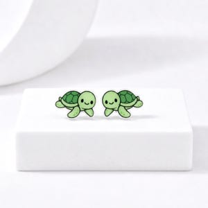 May include: Two pairs of green turtle-shaped earrings. Each earring features a smiling turtle with a green shell and a white background. The earrings are on a white surface.