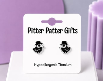 May include: A pair of black and white plague doctor bird stud earrings on a white display card labeled Pitter Patter Gifts and Hypoallergenic Titanium. The earrings feature a stylized bird wearing a top hat and a long beak mask.
