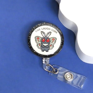 May include: A retractable badge reel with a black glitter border and a white center featuring a cartoon moth with red eyes and the words "LIVE. LAUGH. LURK." The reel has a clear plastic strap and a metal clip.