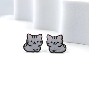 May include: A pair of gray cat-shaped stud earrings. Each earring features a cartoon cat design with black outlines, gray stripes, and pink inner ears. The earrings are set against a white background.