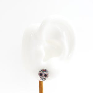 Pu&ograve; includere: Un piccolo orecchino a bottone rotondo con un colorato design a teschio di zucchero. Il teschio ha uno sfondo bianco con dettagli rossi, blu, gialli e neri. L'orecchino &egrave; esposto su una forma bianca a forma di orecchio.