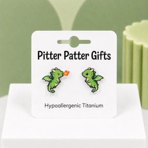 May include: A pair of stud earrings featuring green dragon illustrations on a white display card labeled Pitter Patter Gifts and Hypoallergenic Titanium. One dragon is breathing a small orange flame, while the other is standing with wings spread.