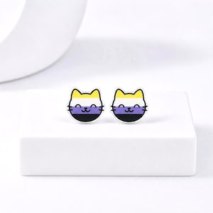 May include: A pair of cat-shaped earrings. Each earring features a yellow, white, purple, and black design, representing the non-binary pride flag. The earrings are set against a white background.