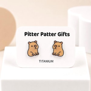 May include: A pair of stud earrings featuring a cartoon capybara design on a white display card labeled Pitter Patter Gifts and TITANIUM. The earrings are light brown with dark brown outlines and facial features, presented on a rectangular card.