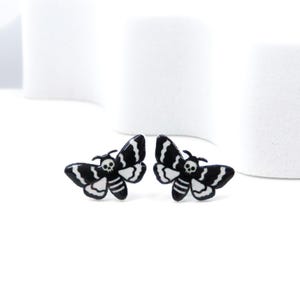 May include: A pair of black and white moth-shaped earrings. Each earring features a skull design on the head and striped wings. The earrings are set against a white background.