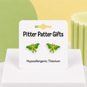 May include: A pair of stud earrings shaped like green luna moths with yellow accents, displayed on a white card labeled Pitter Patter Gifts and Hypoallergenic Titanium. The earrings feature detailed wing patterns and are made of titanium.