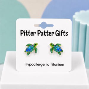 May include: A pair of stud earrings shaped like sea turtles with blue and green enamel detailing and gold-tone outlines. The earrings are attached to a white display card labeled Pitter Patter Gifts and Hypoallergenic Titanium.