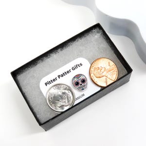 Pu&ograve; includere: Una scatola regalo nera contiene una moneta da dieci centesimi d'argento, un penny di rame e una piccola spilla a forma di teschio di zucchero colorata. La spilla &egrave; circondata da una carta bianca con il testo "Pitter Patter Gifts". La scatola &egrave; foderata con imbottitura bianca.