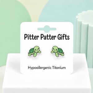 May include: A pair of hypoallergenic titanium stud earrings shaped like small green turtles with dark green shells. The earrings are displayed on a white card featuring the text Pitter Patter Gifts and Hypoallergenic Titanium in black font.