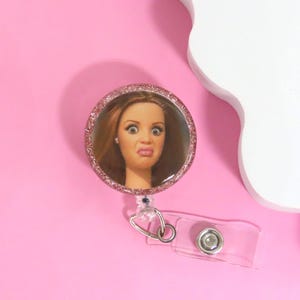 Puede incluir: Un portatarjetas redondo con un borde rosa brillante, que muestra la cara de una mujer con una expresi&oacute;n de sorpresa. El carrete tiene una correa de pl&aacute;stico transparente y un clip de metal, sobre un fondo rosa.