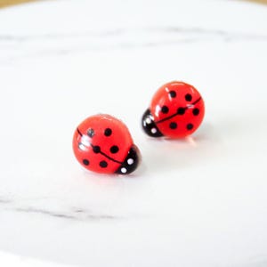 May include: Two small, red ladybug-shaped objects with black spots and black and white heads. The ladybugs are on a white surface. The ladybugs are likely earrings or small decorative items.