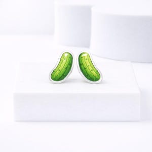 May include: A pair of novelty stud earrings featuring a green pickle design with a silver border. The earrings have a glossy finish and are set against a white background. The earrings are a fun and quirky accessory.