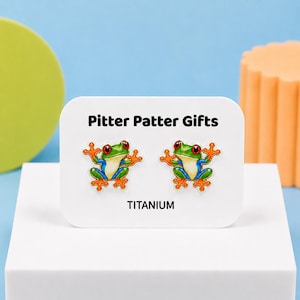 May include: A pair of stud earrings featuring a colorful red-eyed tree frog design with green, blue, and orange details. The earrings are displayed on a white card labeled Pitter Patter Gifts and TITANIUM, indicating the metal material used.