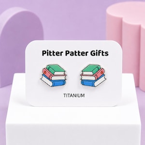 May include: A pair of stud earrings featuring a stack of three books in green, pink, and blue with a small red heart detail. The earrings are displayed on a white rectangular card labeled Pitter Patter Gifts and TITANIUM in black text.