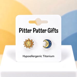 May include: A pair of stud earrings on a white display card labeled Pitter Patter Gifts and Hypoallergenic Titanium. One earring features a yellow sun with a face, and the other is a blue crescent moon with a face and yellow stars.