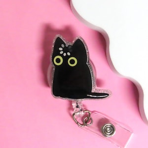 May include: A black cat-shaped badge reel with glitter trim and yellow eyes. The retractable reel has a clear plastic strap and a metal clip. The background is pink with a white cloud-shaped design.