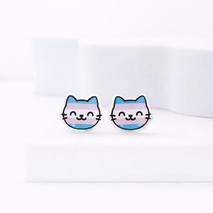 May include: A pair of cat-shaped stud earrings. Each earring features a smiling cat face with a blue, pink, and white striped design, representing the transgender pride flag. The earrings have a silver-colored border and a simple, clean design.