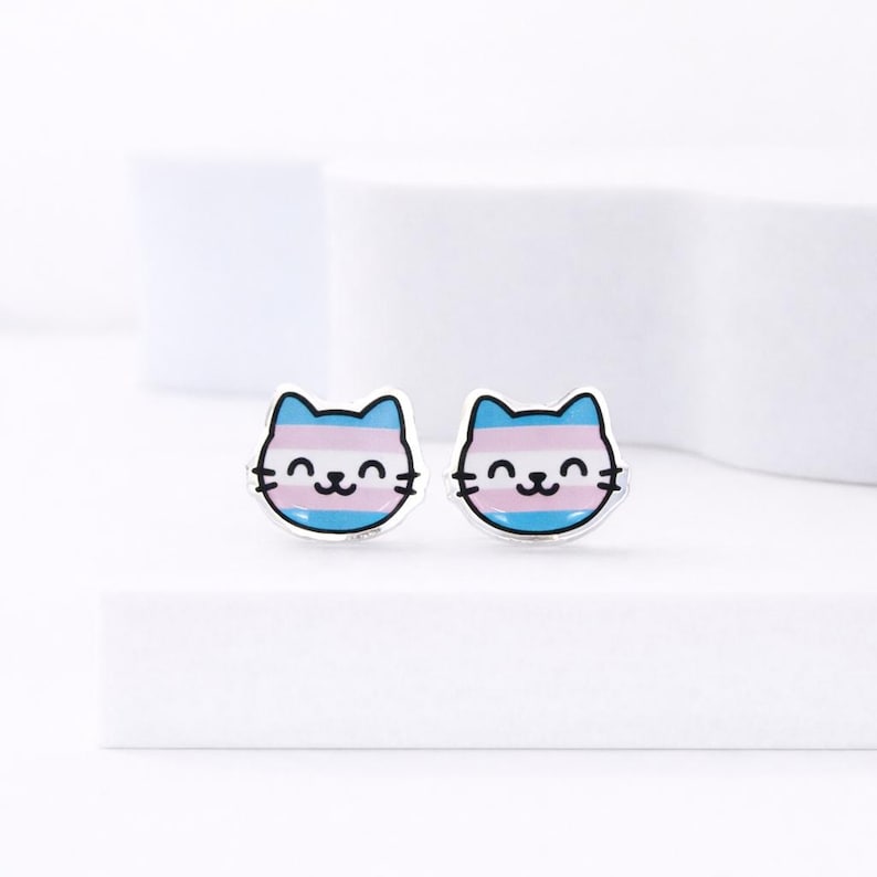 May include: A pair of cat-shaped stud earrings. Each earring features a smiling cat face with a blue, pink, and white striped design, representing the transgender pride flag. The earrings have a silver-colored border and a simple, clean design.