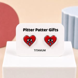 May include: A pair of heart-shaped stud earrings on a white display card labeled Pitter Patter Gifts and TITANIUM. Each red heart features a central eye with long black lashes and three blue teardrops falling from the eye, rendered in a graphic style.