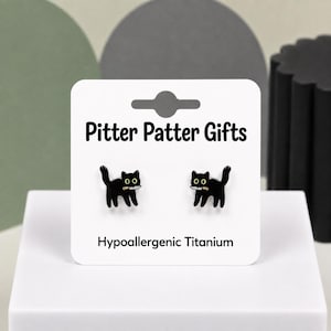May include: A pair of black cat stud earrings with white knives in their mouths, displayed on a white card labeled Pitter Patter Gifts and Hypoallergenic Titanium. The cats have yellow eyes and are depicted in a standing, side-profile position.