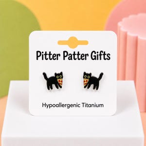 May include: A pair of black cat stud earrings, each featuring a cat holding a slice of pepperoni pizza. The earrings are displayed on a white card labeled Pitter Patter Gifts and Hypoallergenic Titanium, with a cutout at the top for hanging.