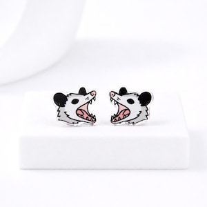 May include: A pair of opossum-shaped stud earrings. Each earring features a white opossum head with black ears, a pink mouth, and sharp teeth. The earrings are set against a white background.